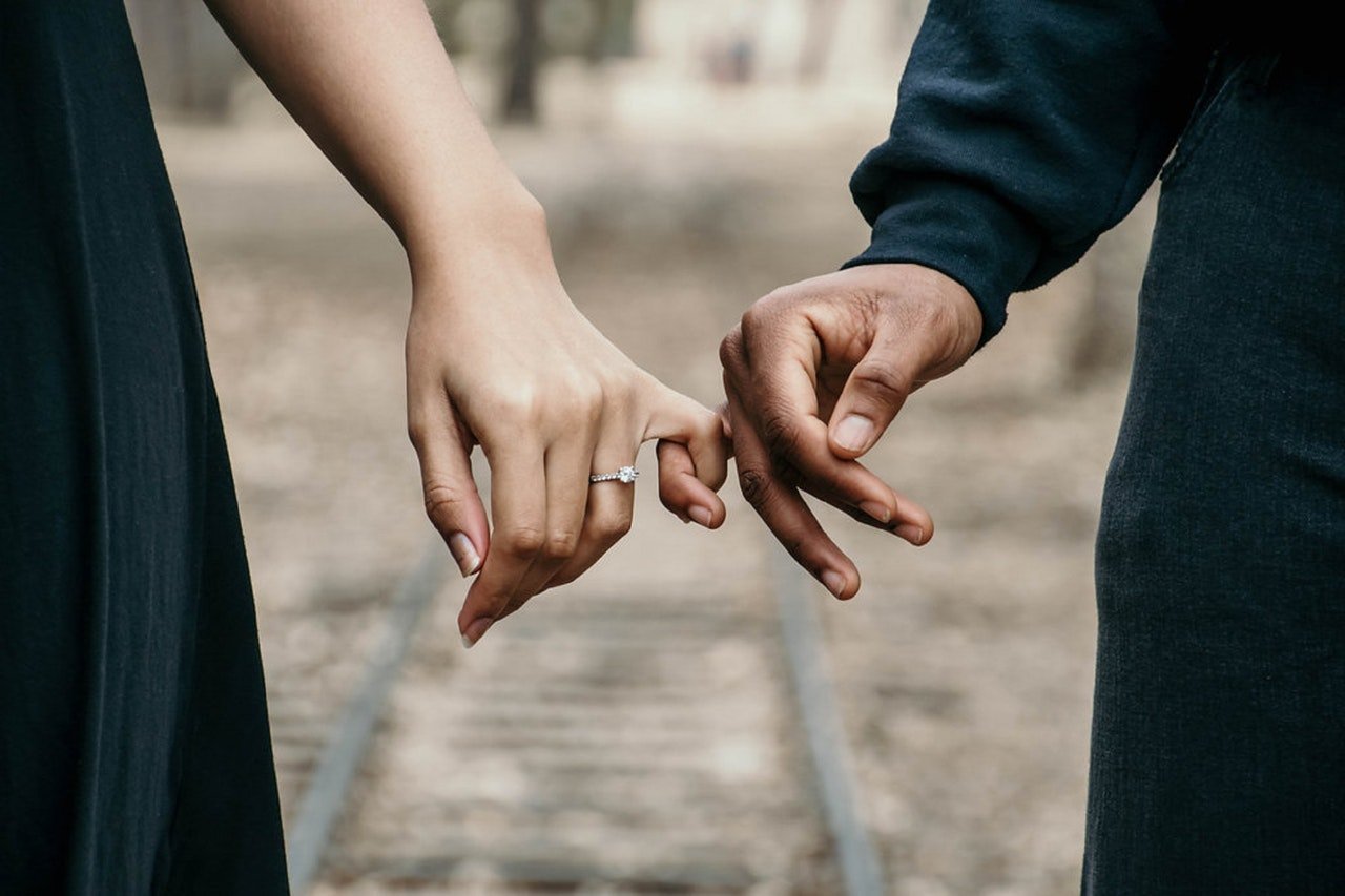 Homem e mulher com dedos mindinhos entrelaçados. A mulher tem um anel de noivado no dedo anelar.