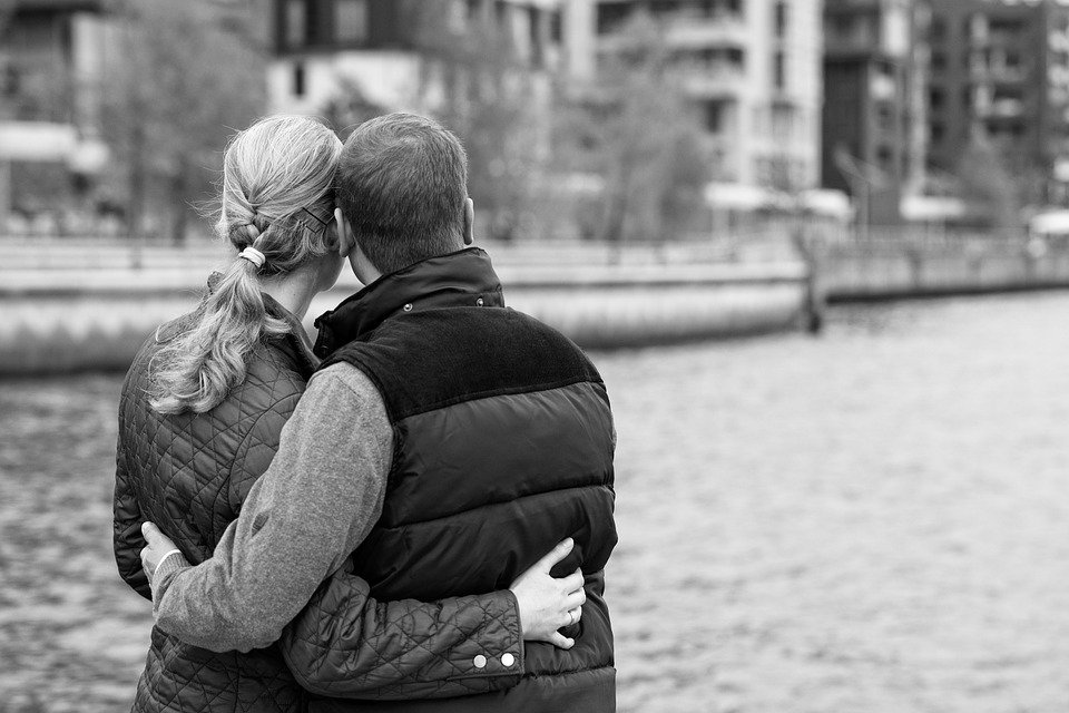 Mulher e homem abraçados olhando para um rio canalizado com uma cidade ao lado.