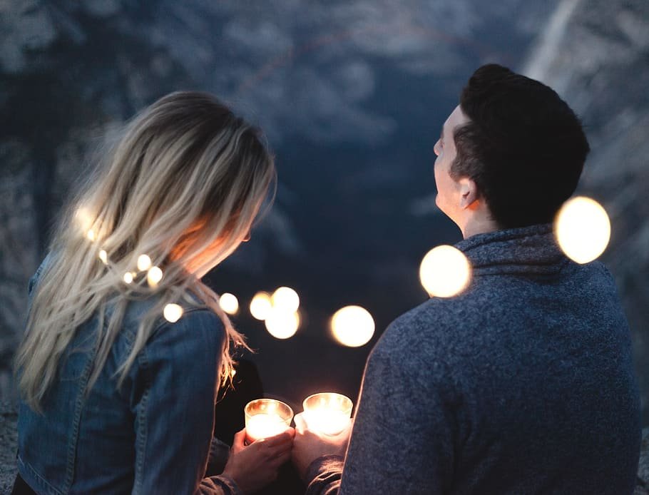 Mulher e homem com luzes ao redor do pescoço, sentados em uma montanha, segurando copos com velas.