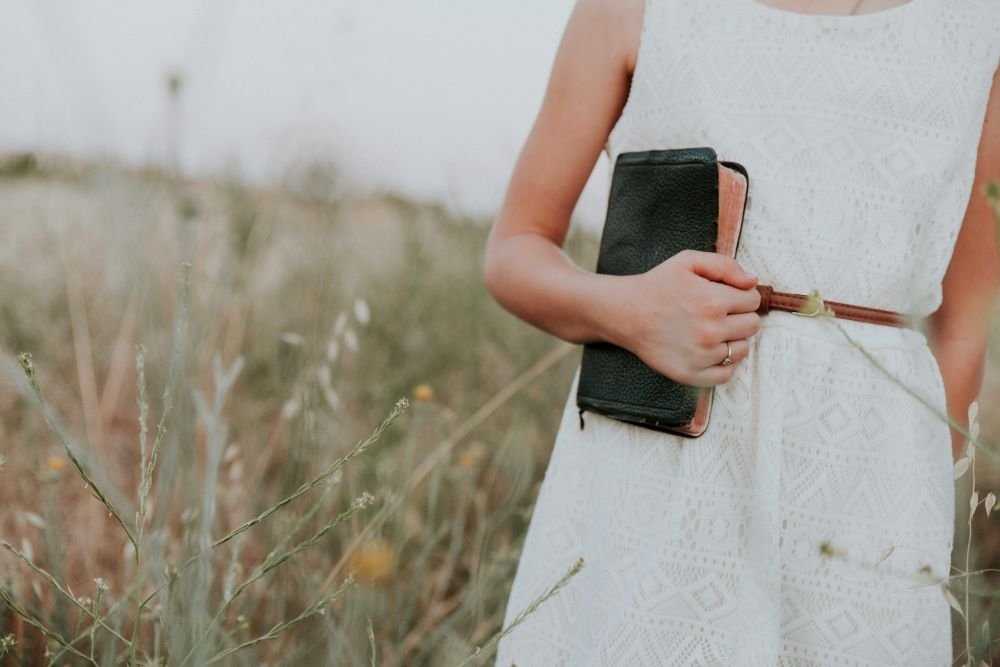 Mulher carregando a bíblia em seu braço andando por um campo de flores.