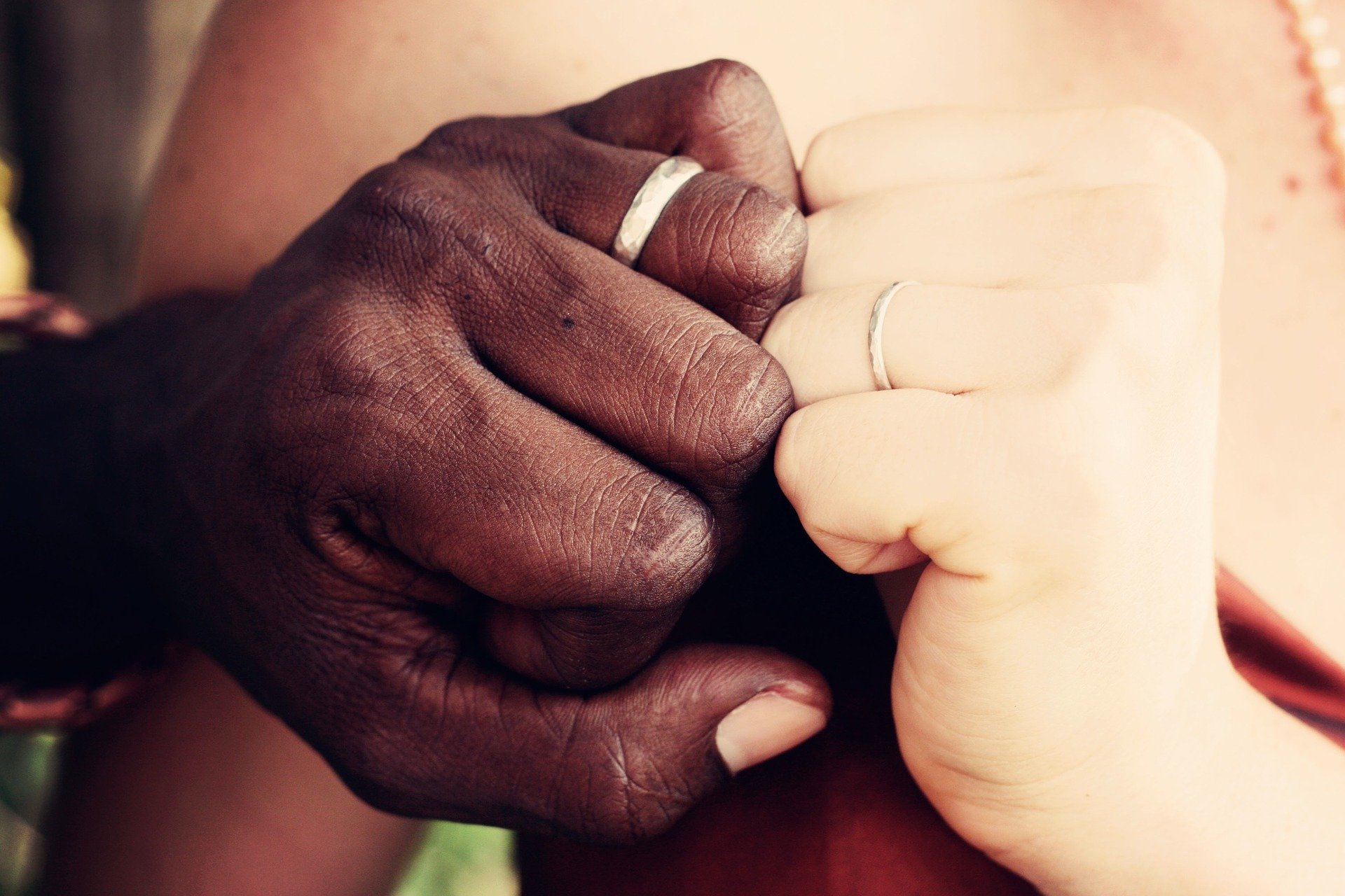 Foto de mãos, uma feminina branca e uma masculina negra, mostrando alianças de noivado