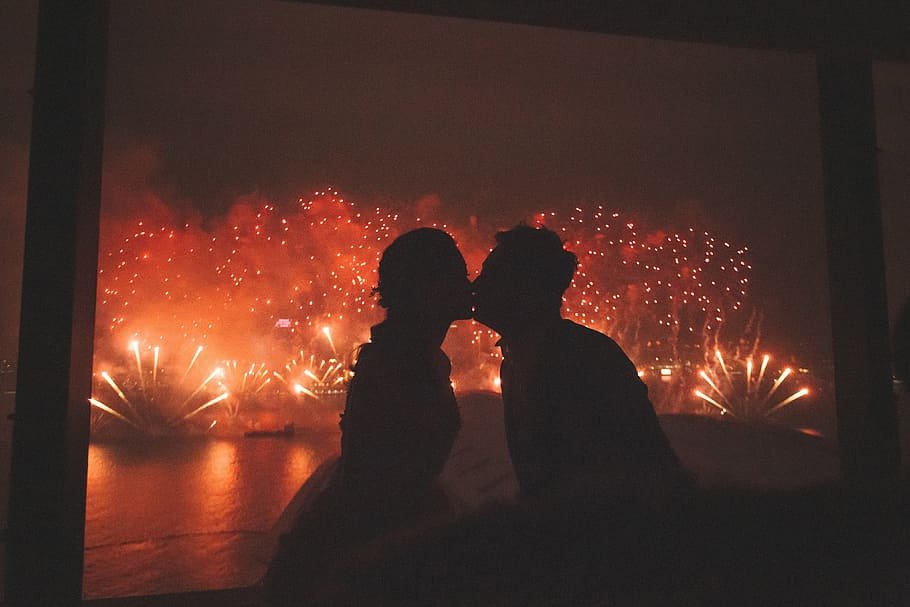 Silhueta de casal se beijando com fogos de artifício cobrindo o céu ao fundo.