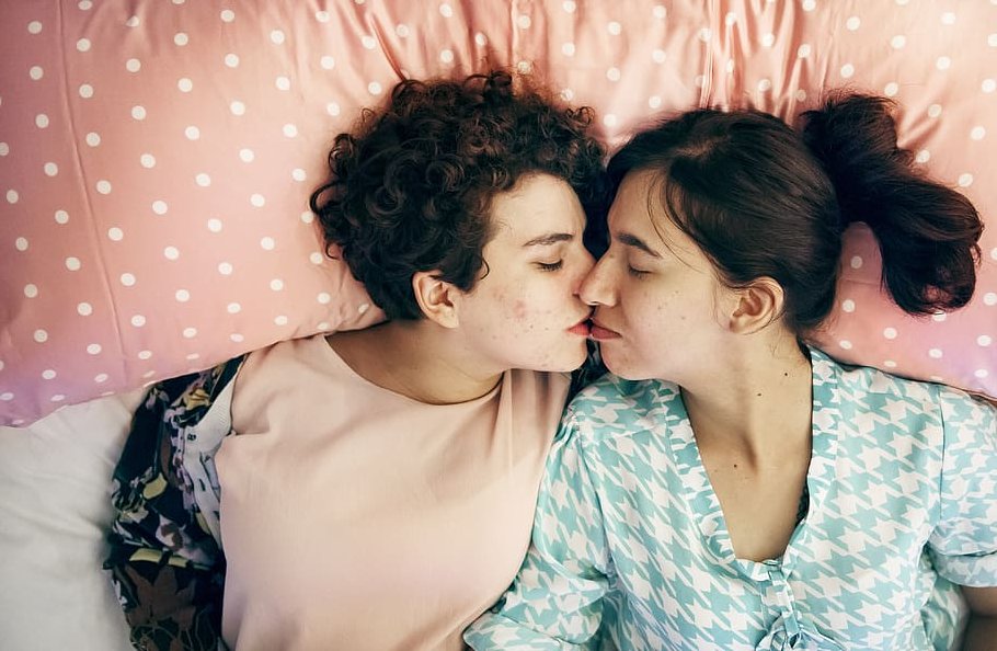 Duas mulheres deitadas com a barriga para cima numa cama, lado a lado. Os rostos delas estão virados para o lado e seus lábios quase se tocam.