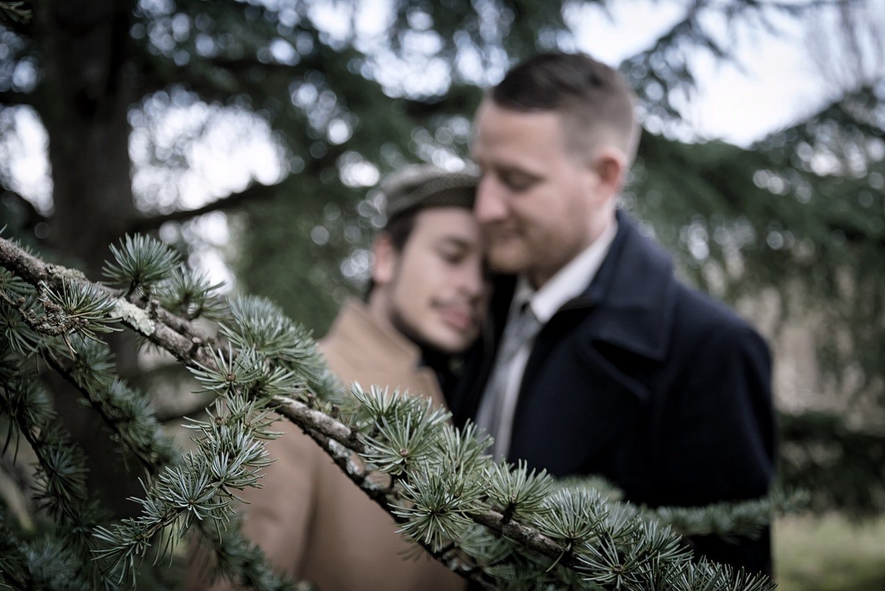 Casal de dois homens abraçados em meio a árvores de uma paisagem natural.