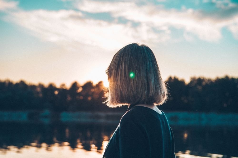 Mulher parada diante de um lago encarando o pôr do Sol.