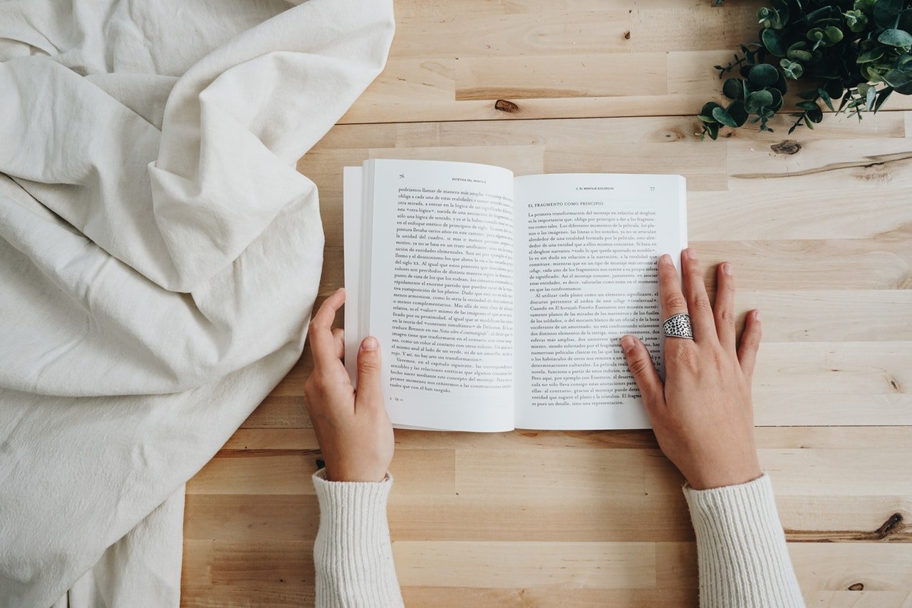 Pexels/Esther Muñoz Trilla Mãos de uma pessoa sobre um livro aberto, em uma mesa de madeira.
