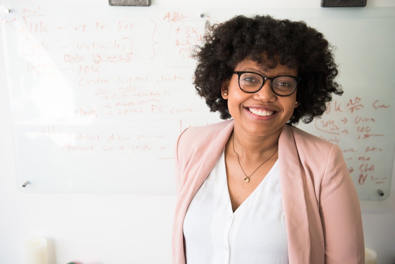 Professora universitária em frente a um quadro branco com escritos, sorrindo.