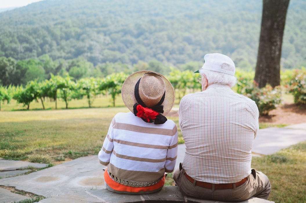 Homem e mulher idosos sentados em um banco, de costas, olhando uma paisagem natural do campo.
