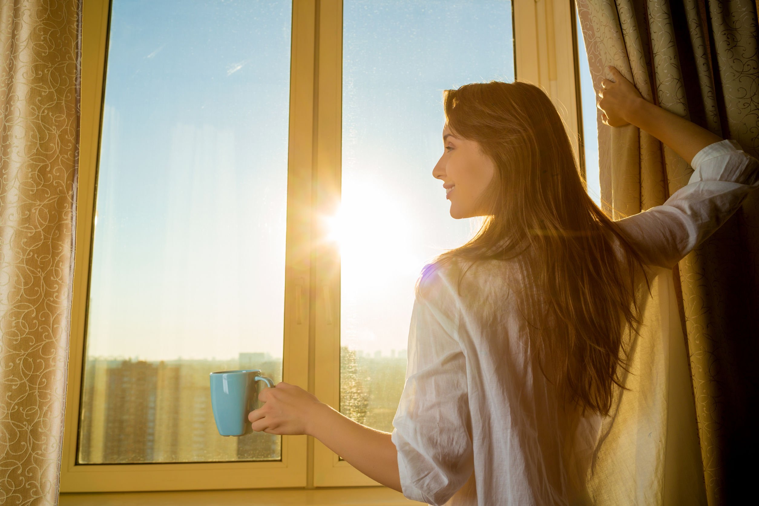 Mulher segurando uma cortina com a mão direita e segurando uma caneca com a mão esquerda. Ela olha pela janela, que está muito clara pela luz do sol.
