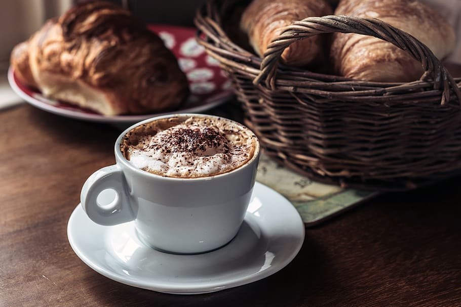 Xícara de chocolate quente, cesta com croissants e um prato com um croissant.