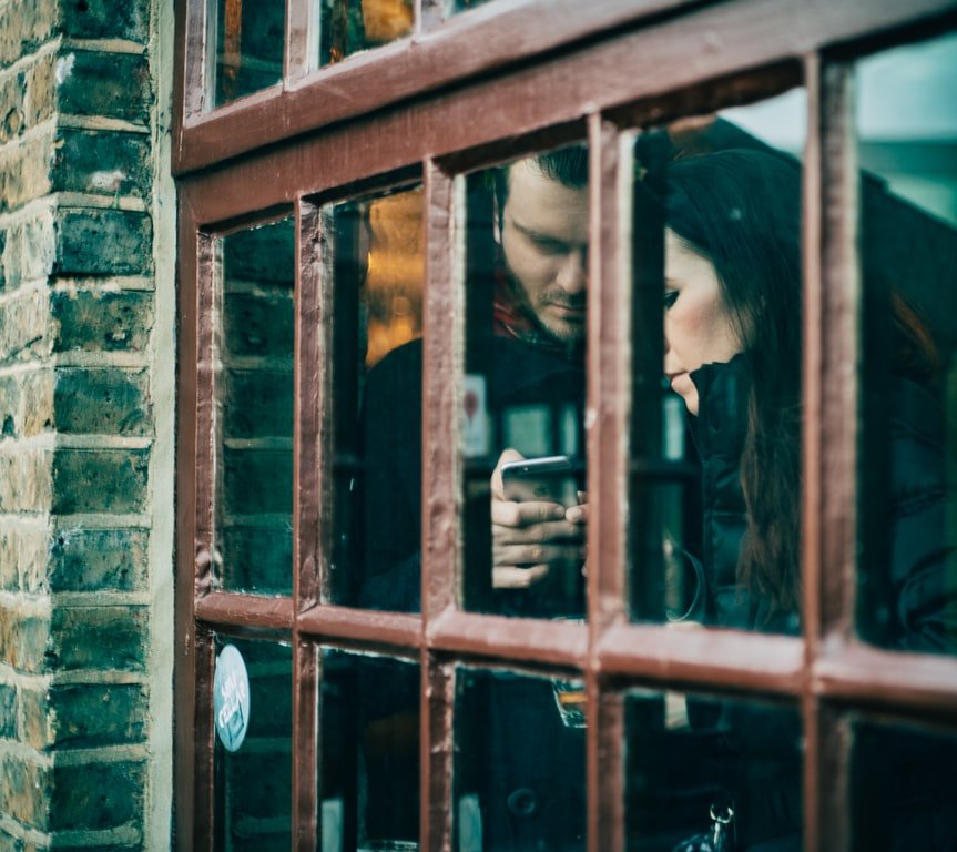 Homem e mulher sérios olhando para o mesmo smartphone, vistos de uma janela.