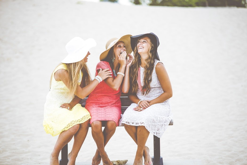 Três mulheres sentadas em um banco na praia, conversando e rindo.