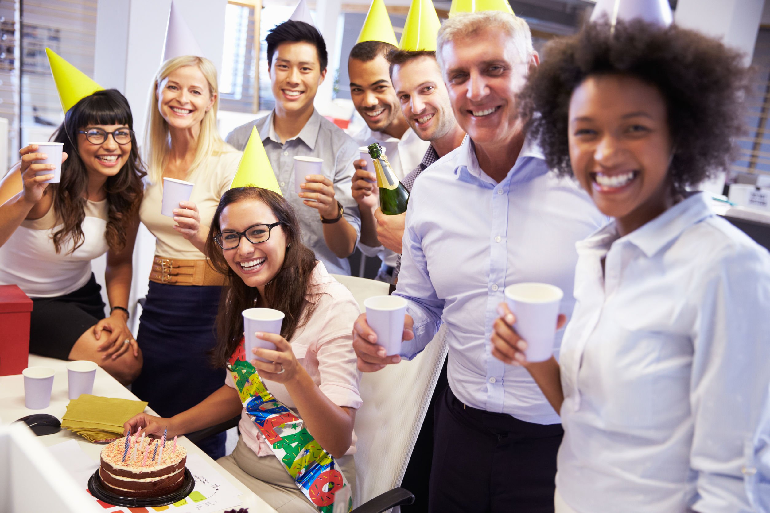 Pessoas comemorando aniversário em escritório posando para foto enquanto seguram copos