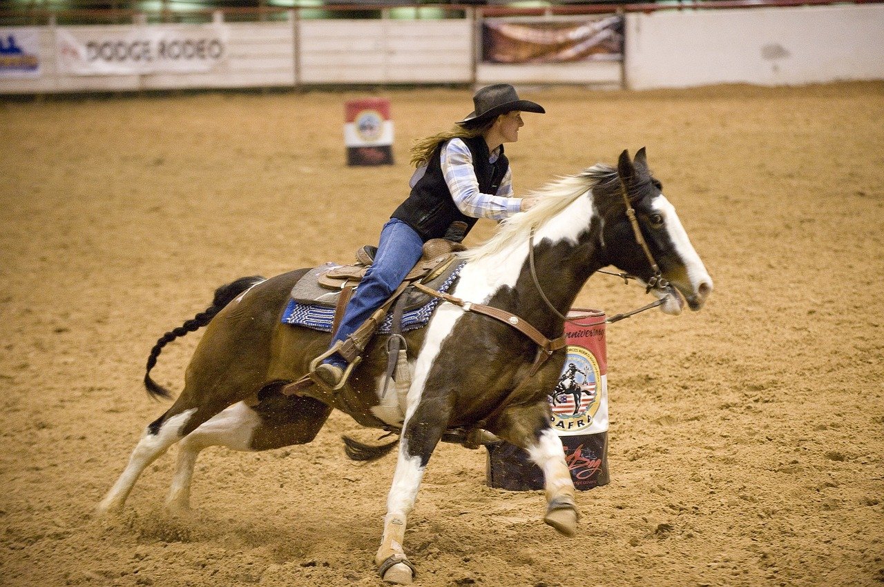Mulher usando chapéu de caubói em cima de cavalo selado, correndo com ele dentro de uma arena.