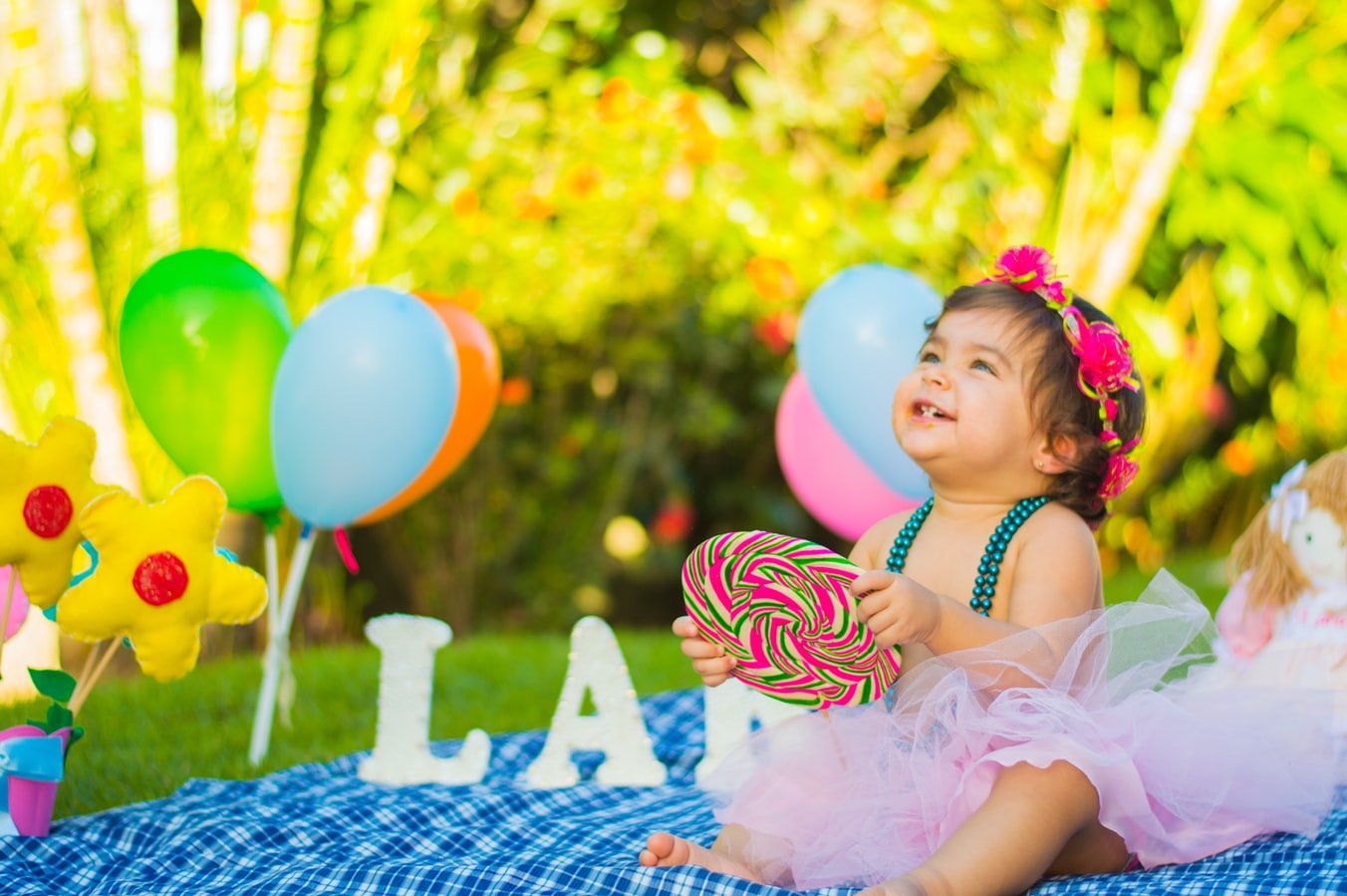 Menina criança usando fantasia, sentada em uma toalha de piquenique, segurando um pirulito em meio a balões de aniversário.