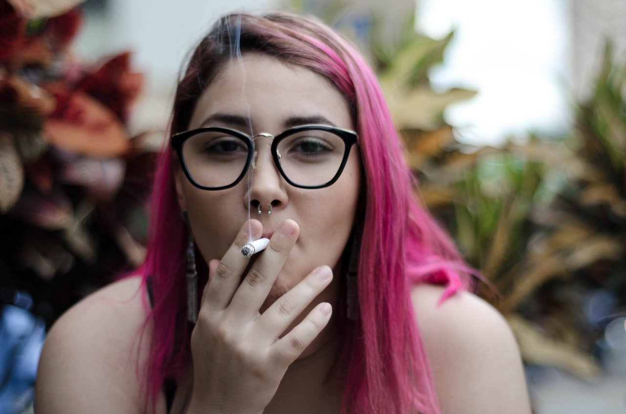 Mulher com cabelo rosa, segurando um cigarro enquanto o fuma.