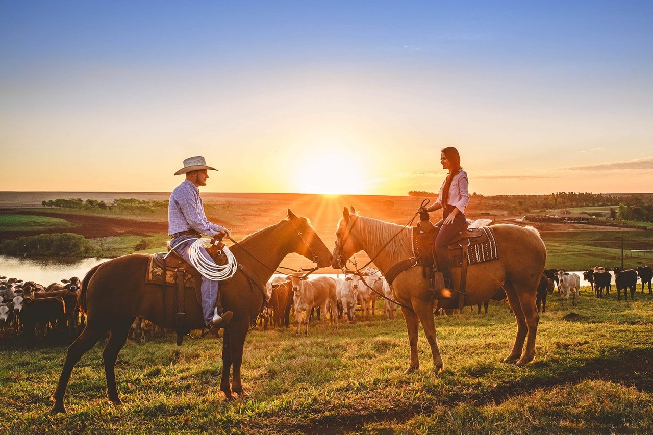 Homem e mulher montados em cavalos, de frente um para o outro, ao pôr do sol.