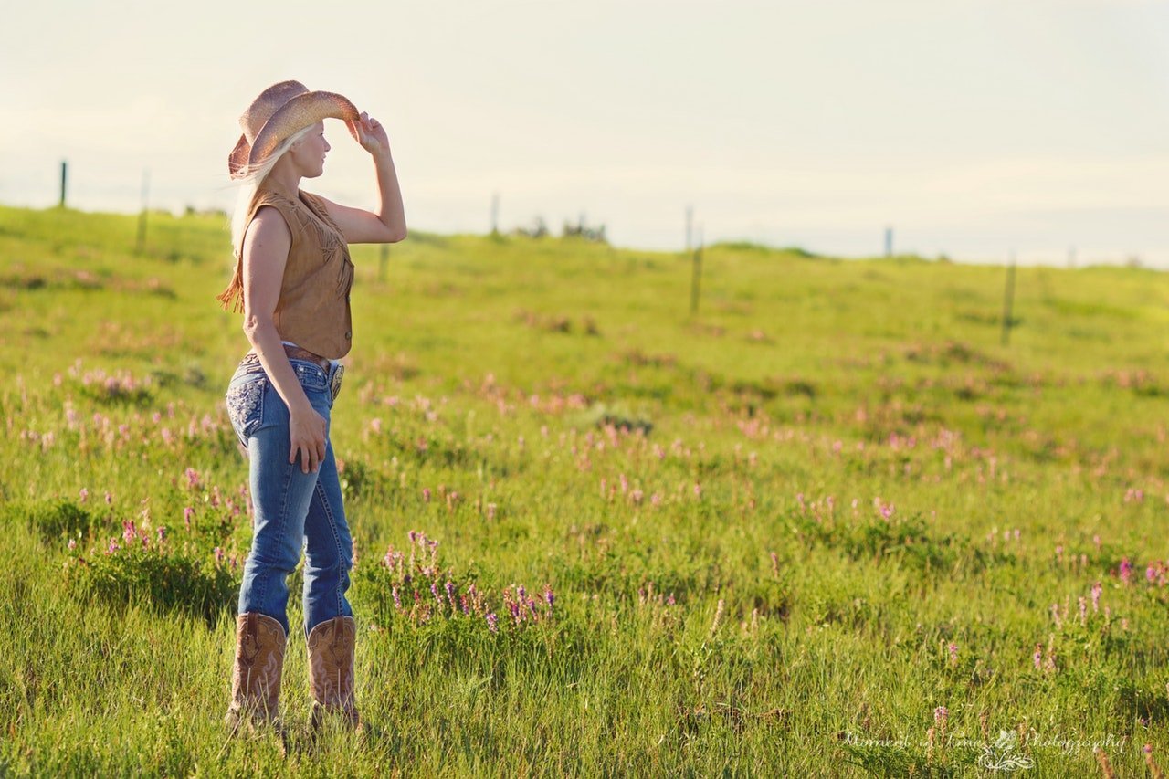Mulher usando roupas de cowgirl no campo, olhando para o lado com a mão no chapéu.