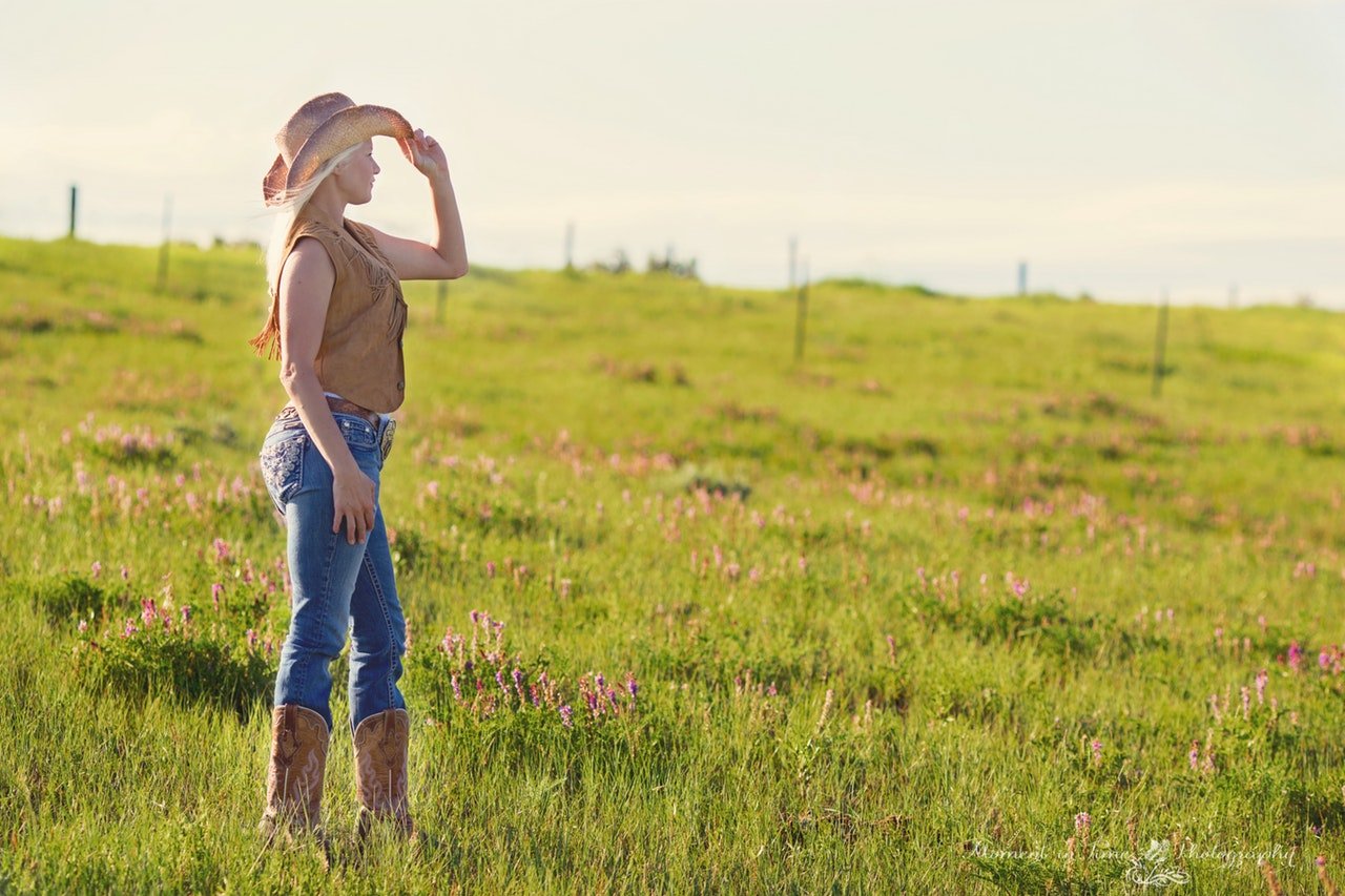 Pixabay Mulher usando roupas de cowgirl no campo, olhando para o lado com a mão no chapéu.