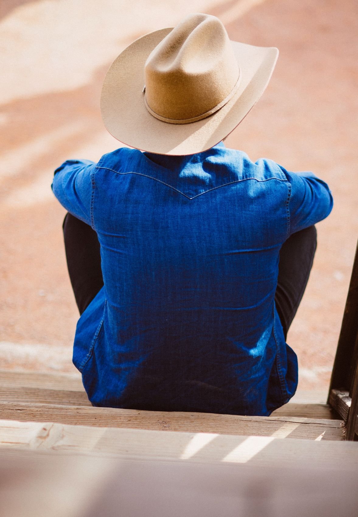 Homem sentado de costas, com camiseta jeans e chapéu de cowboy.