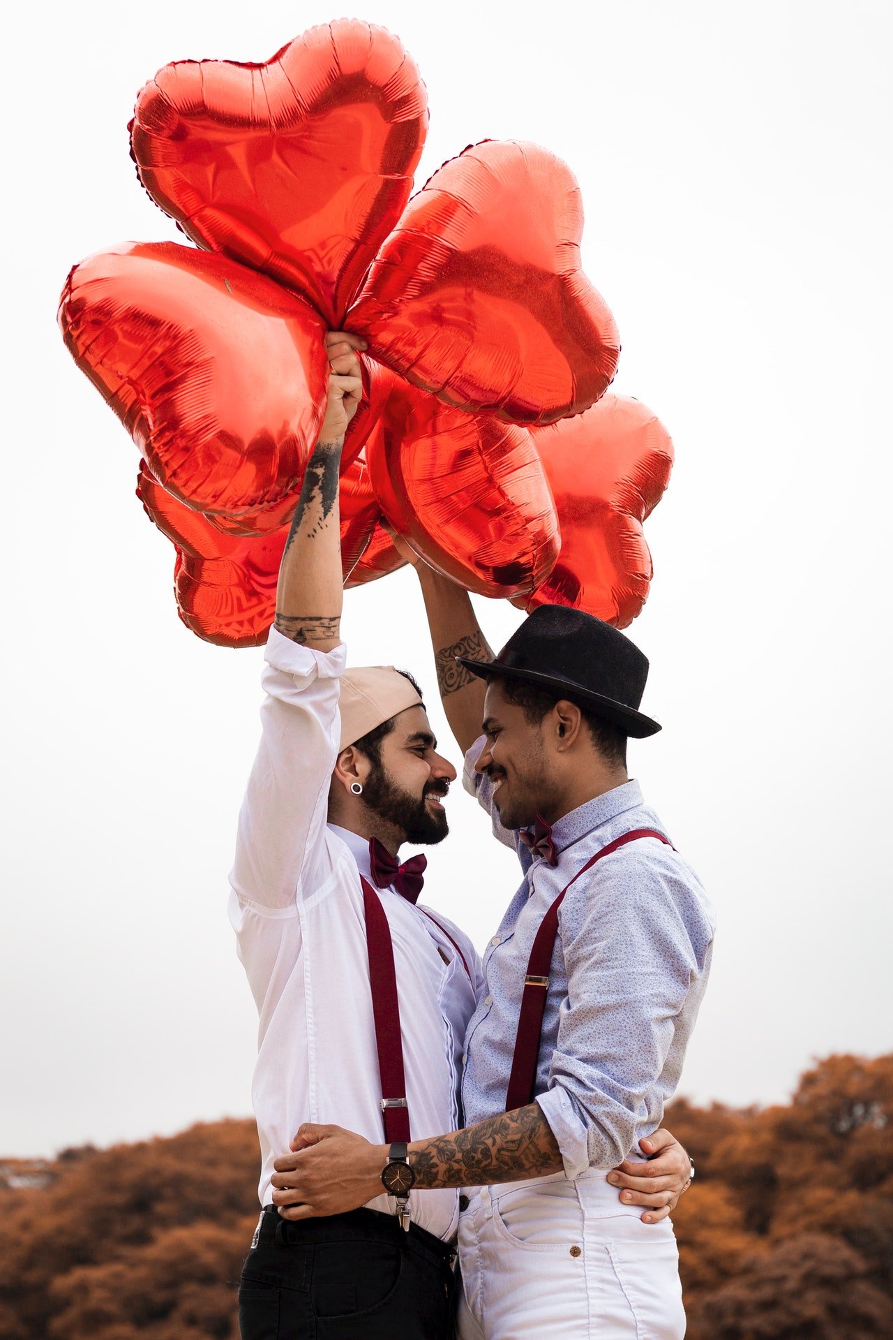 Casal de homens abraçados, segurando balões de coração.