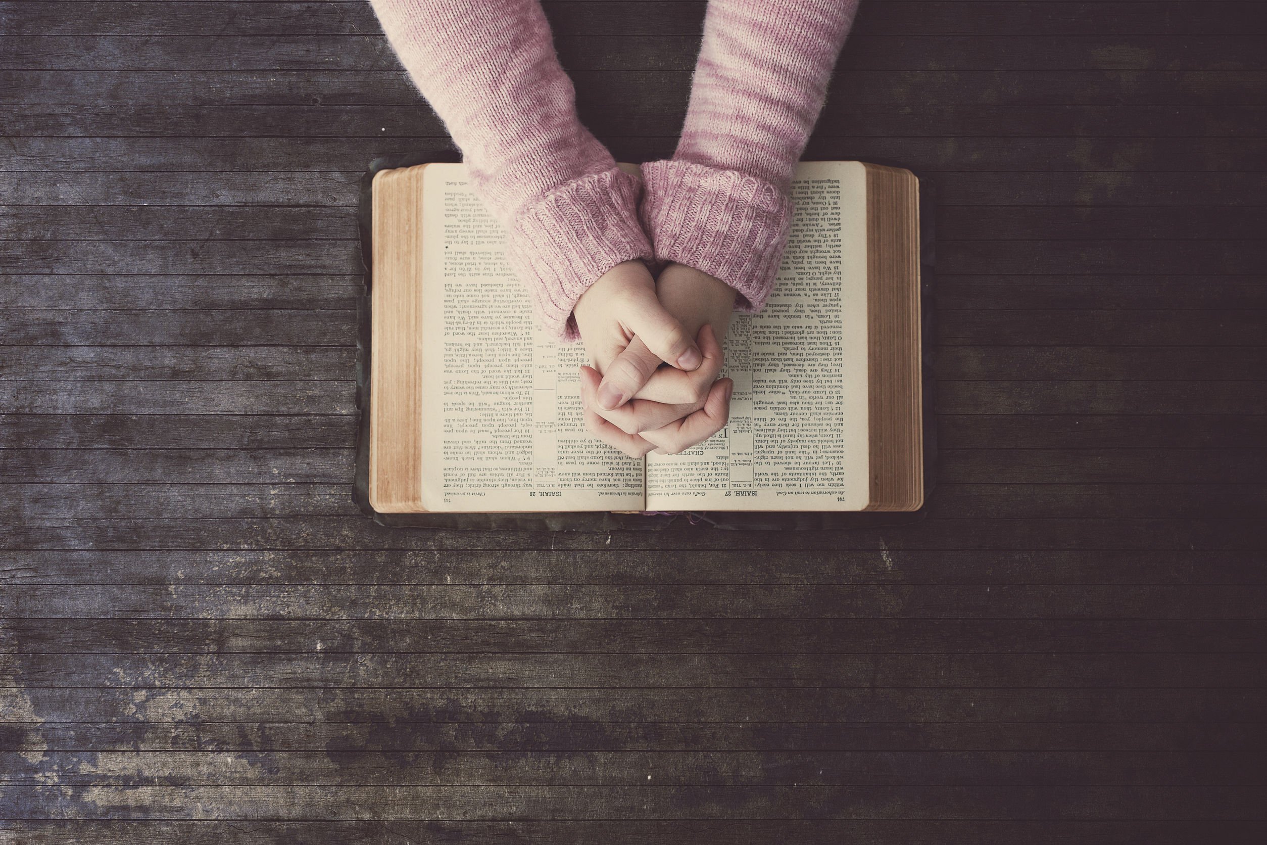 Braços de mulher usando um suéter rosa, com os dedos das duas mãos entrelaçados em cima de uma bíblia aberta.