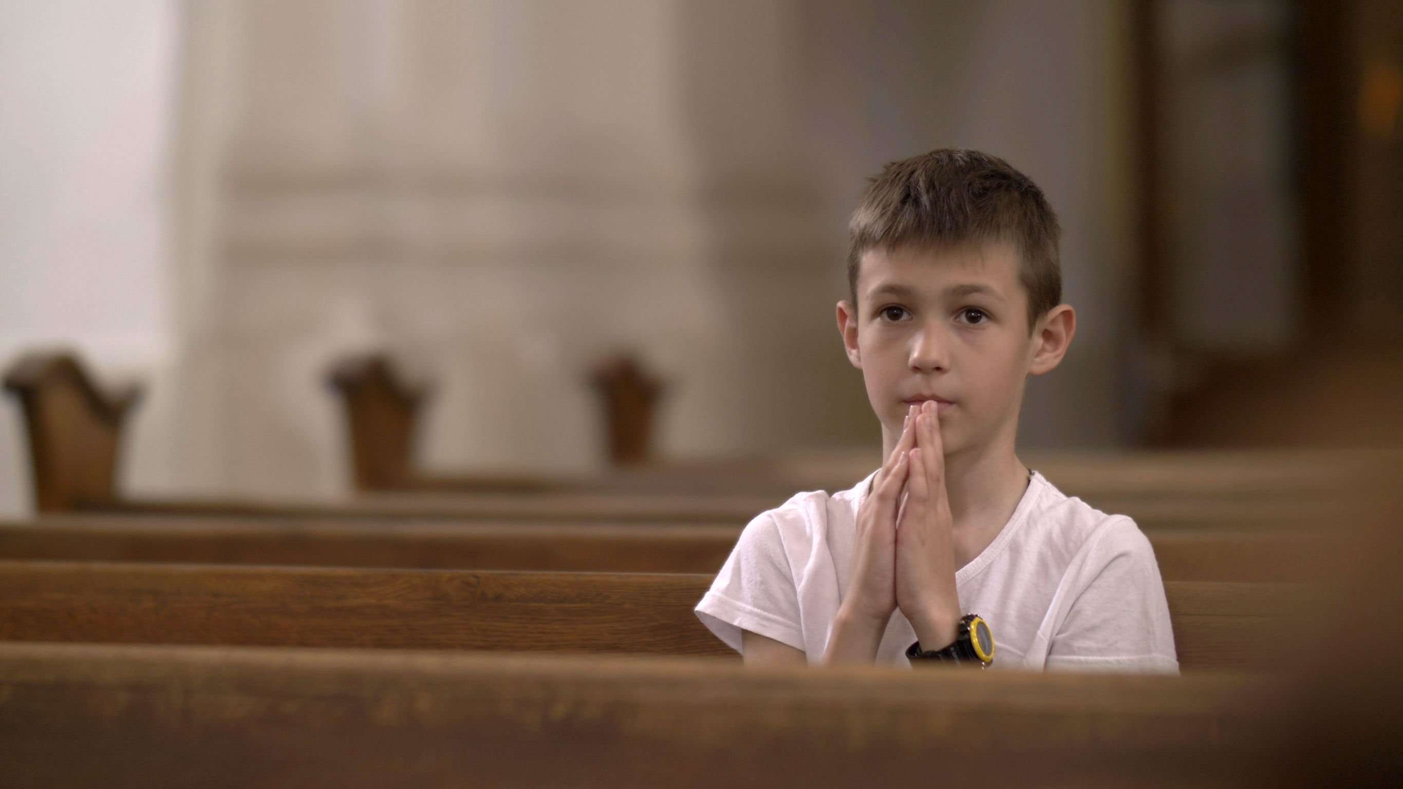 Menino sentado em banco de igreja sozinho, com as palmas das mãos unidas na frente de seu rosto.