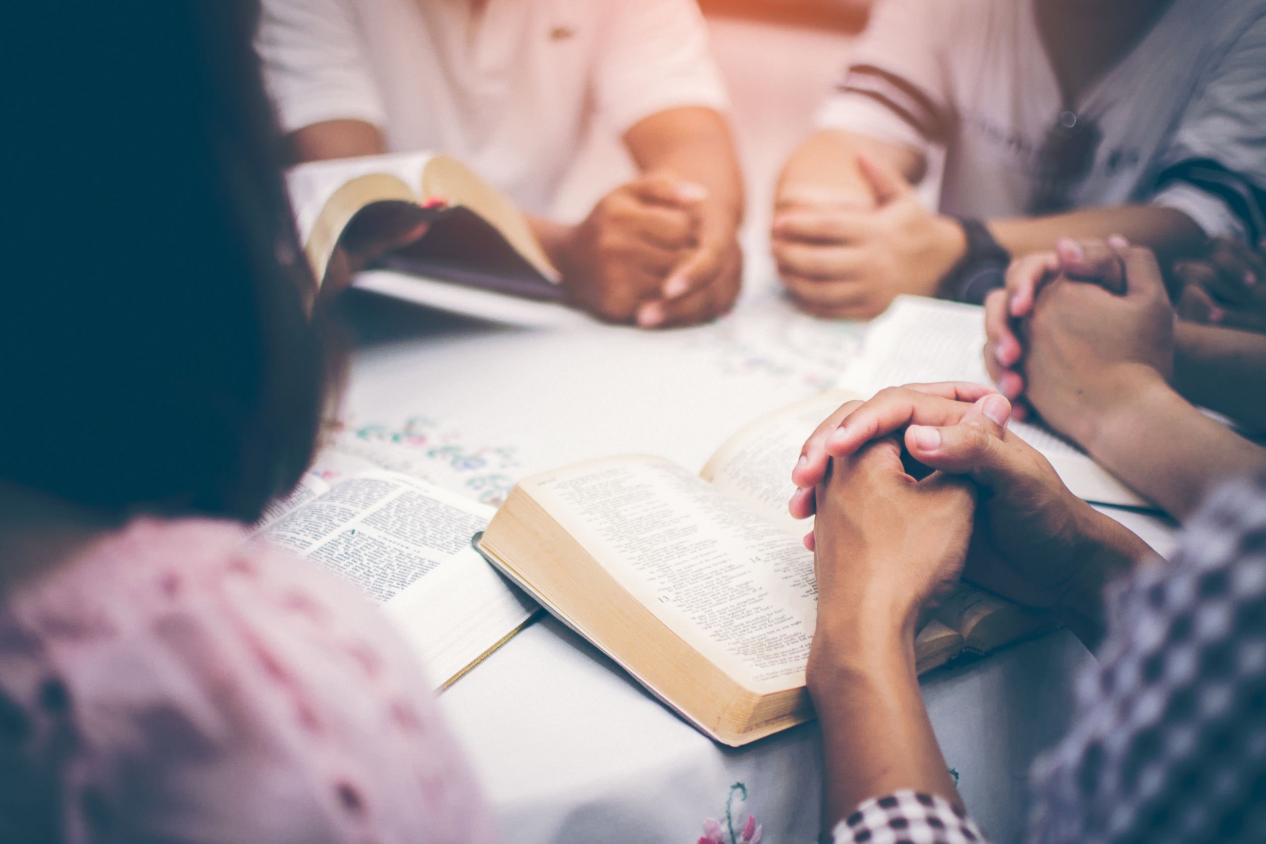 Grupo de pessoas sentado ao redor de uma mesa, cada um com suas mãos entrelaçadas em cima de um exemplar da bíblia.