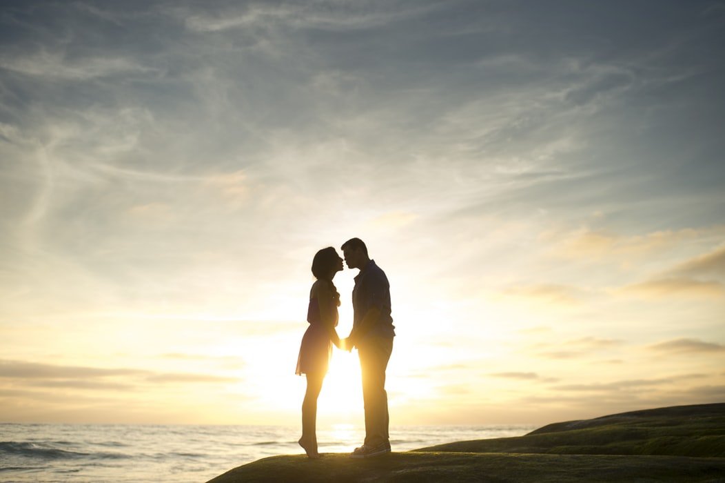 Silhueta de homem e mulher em pequena montanha na beira do mar durante o pôr do sol. Eles estão de mãos dadas e a mulher está na ponta dos pés para beijar o homem.