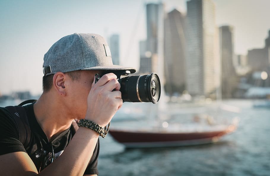 Homem de perfil, usando boné e tirando foto com câmera profissional. Ao fundo, um barco em um rio e muitos prédios.
