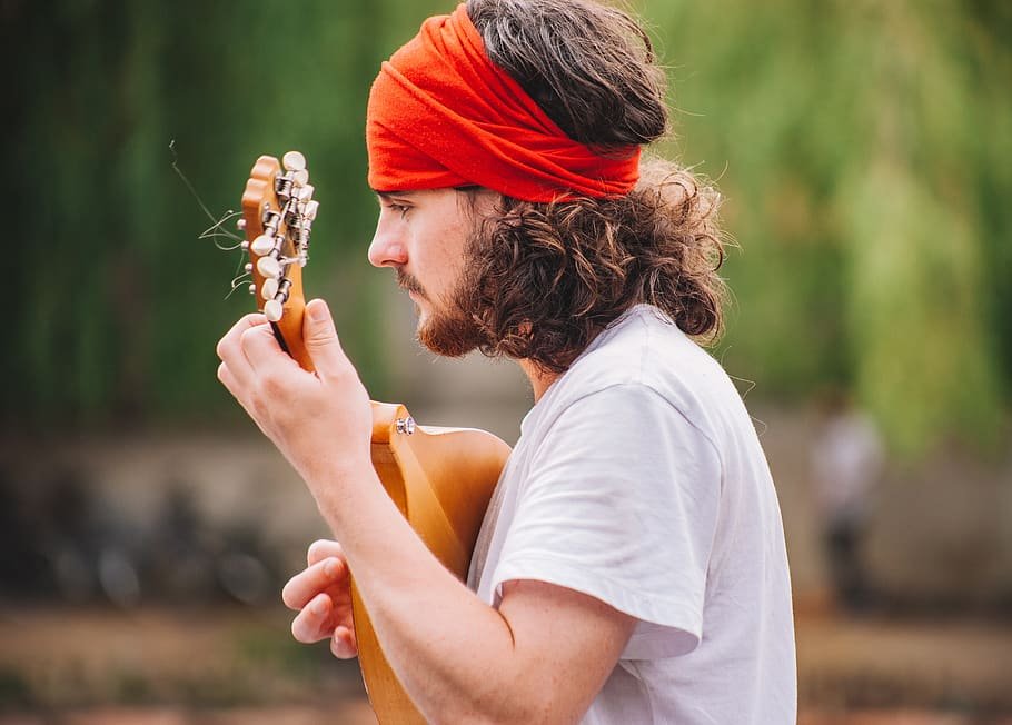 Homem com cabelo cacheado comprido, com uma bandana na testa. Ele está de lado, e tocando um violão.