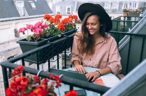 Pexels/Daria Shevtsova Mulher sentada na sacada de um apartamento, rindo, cercada por vasos de flores.
