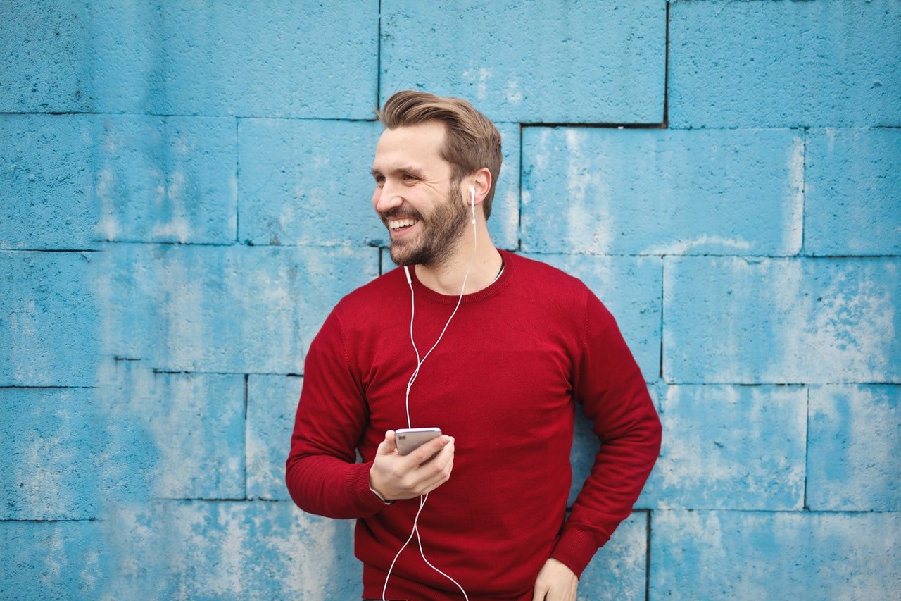 Homem sorrindo em frente a um muro, segurando um smartphone e usando fones de ouvidos.