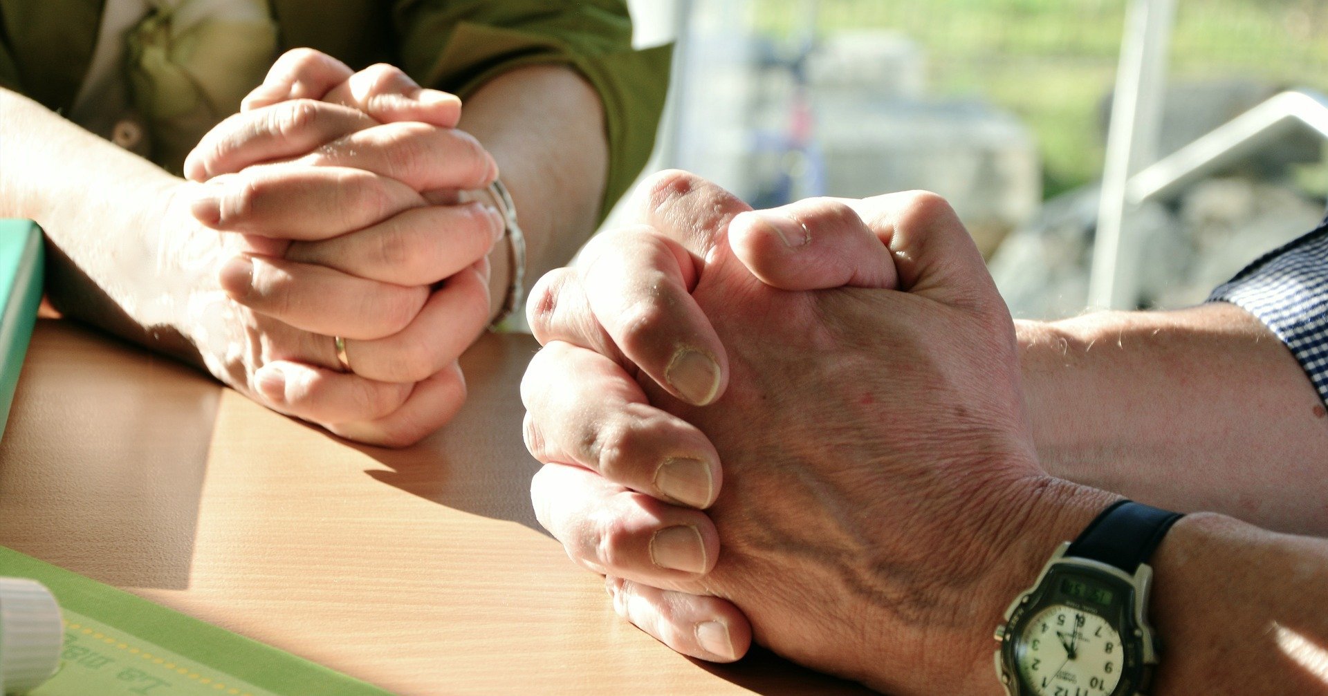 Duas pessoas orando com as mãos fechadas apoiadas em mesa de madeira
