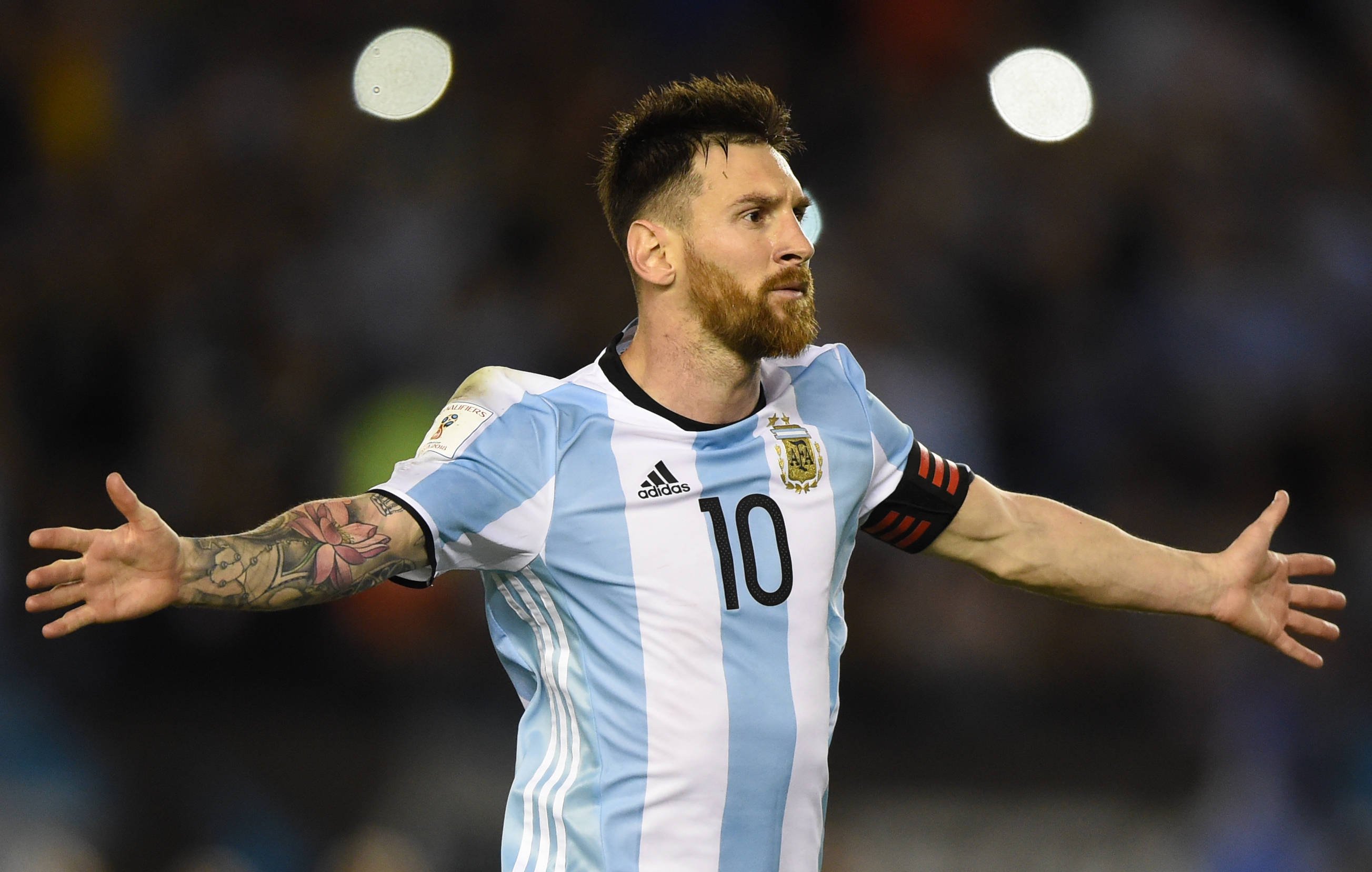 Lionel Messi em campo com o uniforme da Seleção Argentina e os braços abertos.
