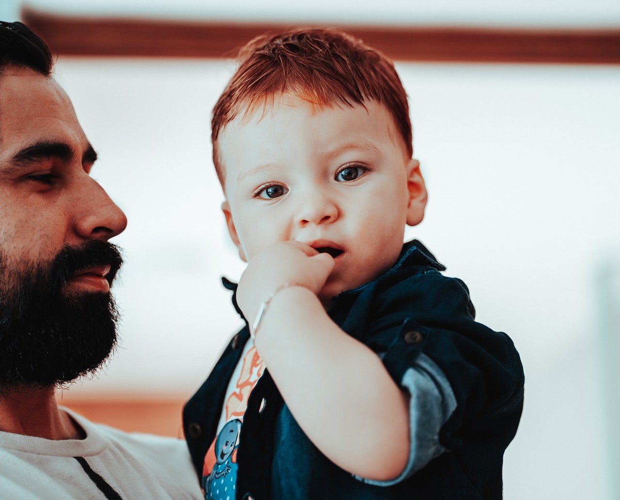 Homem com barba, segurando bebê que olha para a câmera com a mão na boca.
