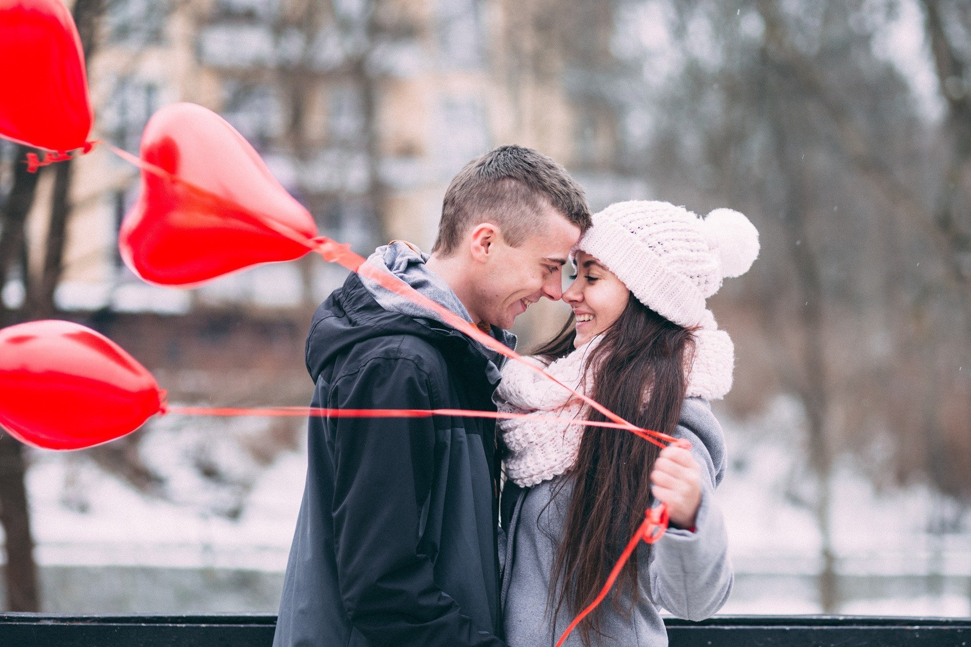 Homem e mulher abraçados sorrindo, e mulher segurando balões em formato de coração.