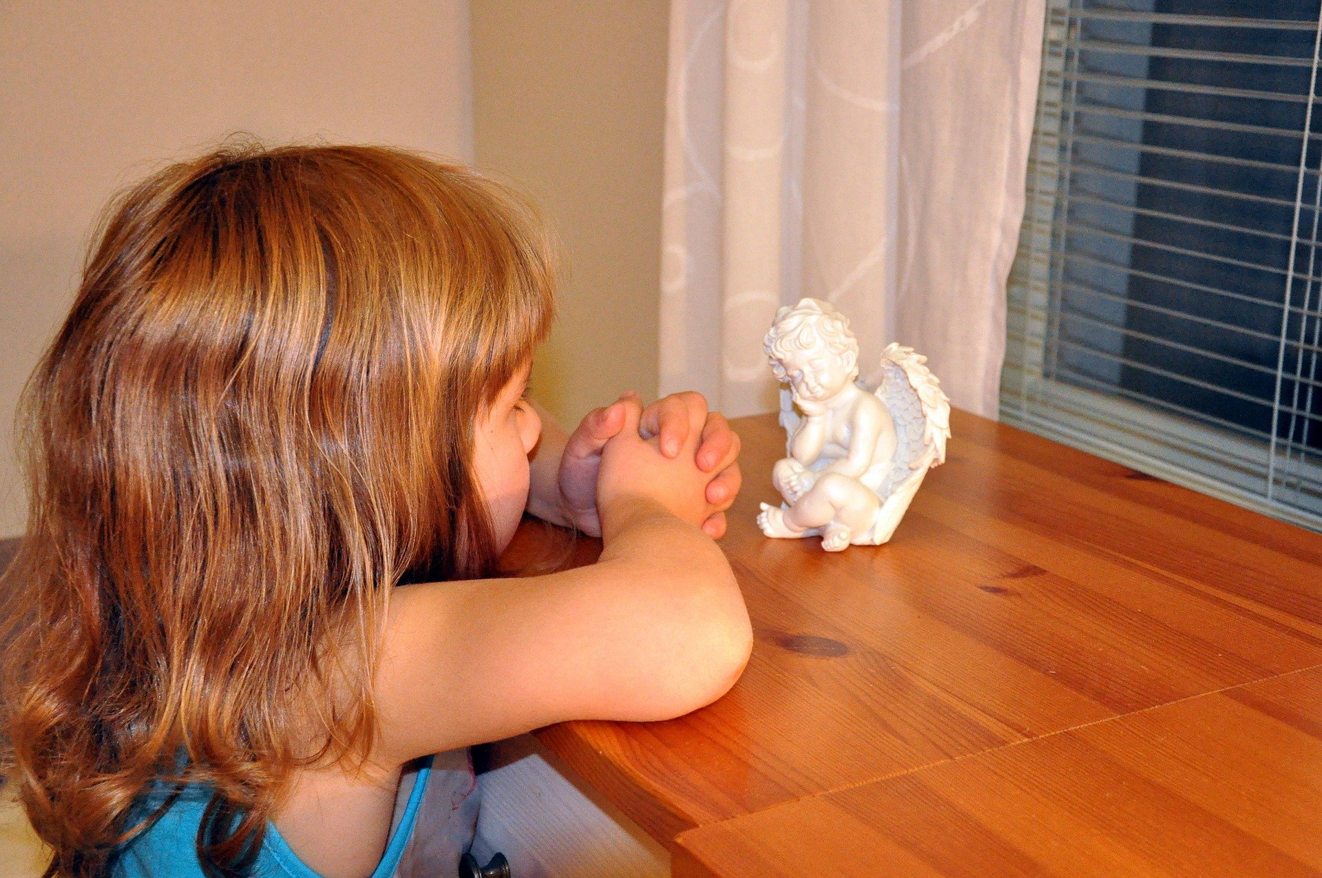 Menina orando debruçada sobre comoda encarando uma estátua branca de anjo