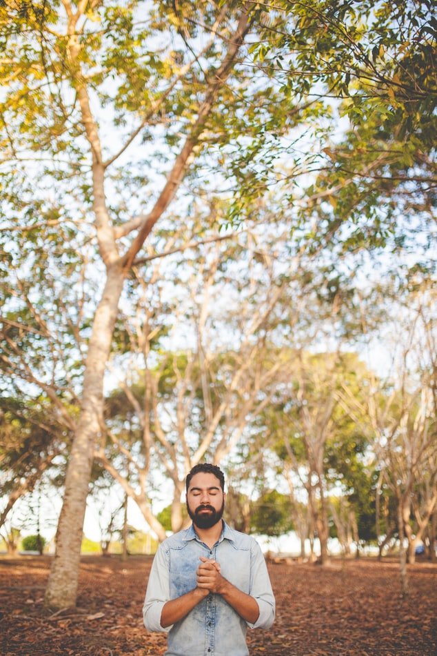 Homem orando de olhos fechados, com as mãos juntas no meio de parque