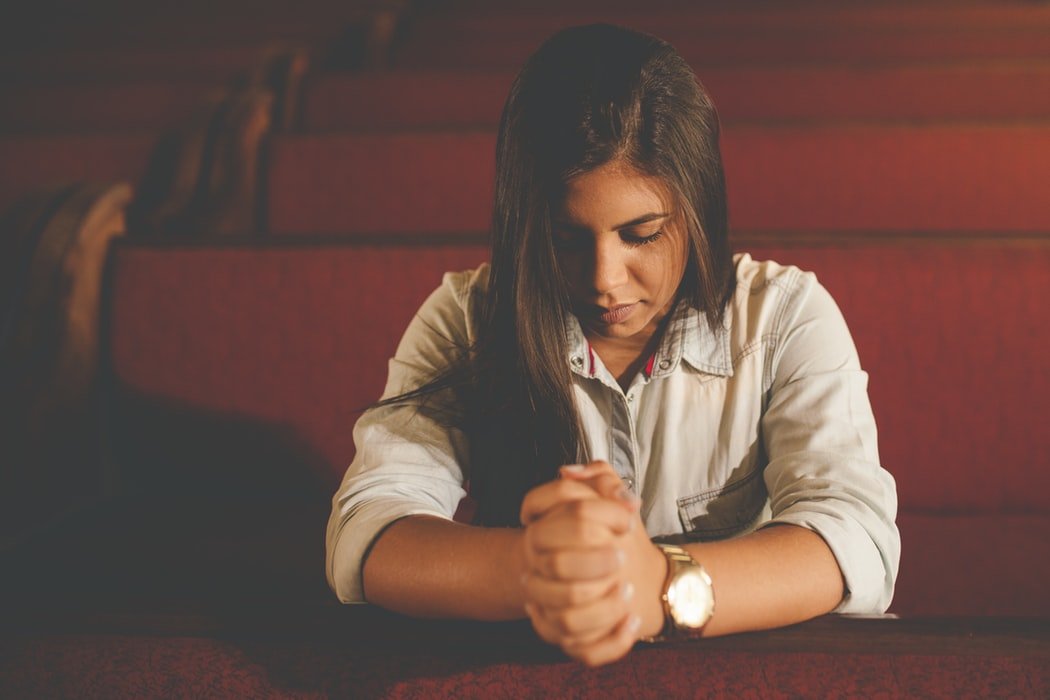 Mulher ornando com as mãos juntas e olhos fechados, sentada em bancos de igreja