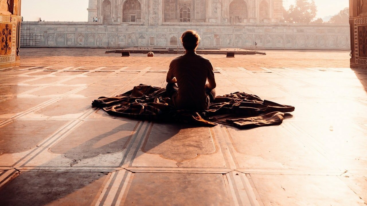 Homem sentado no chão sobre um tecido. Ele está em frente ao Taj Mahal.