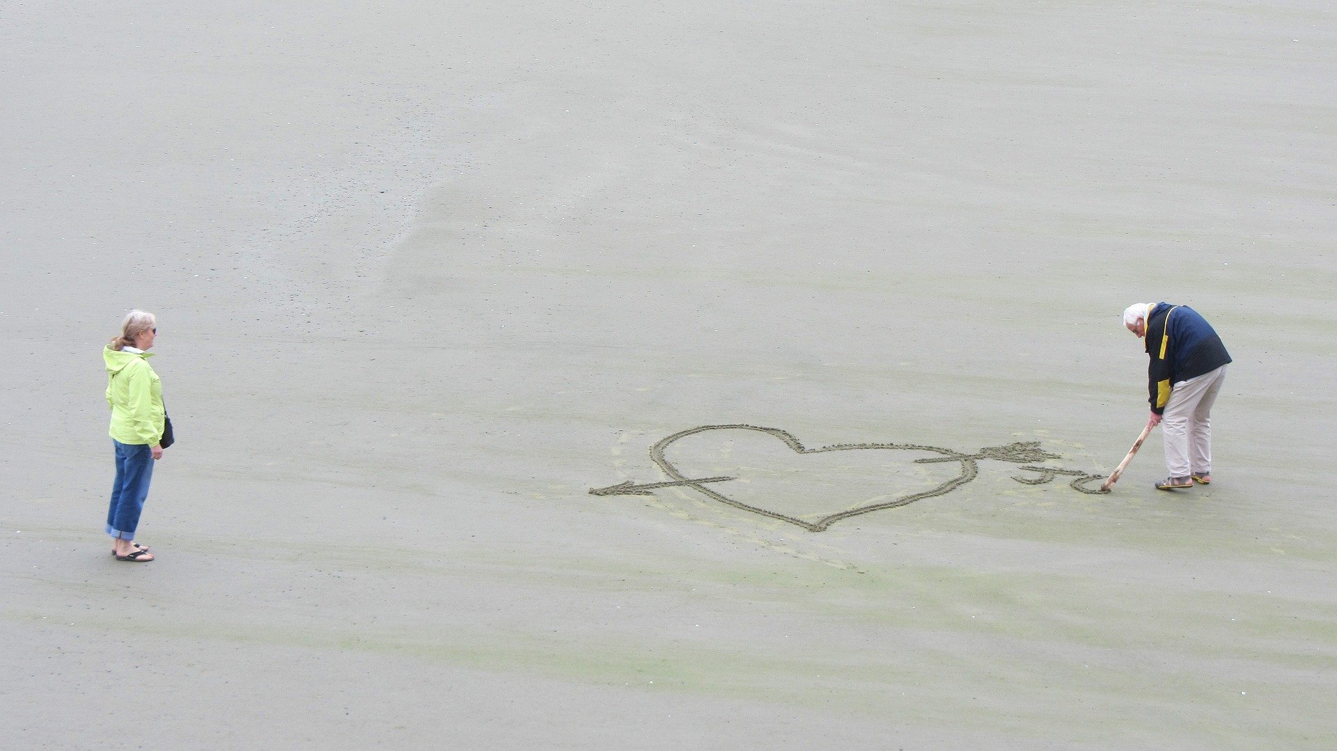 Idoso desenhando coração em areia de praia apontando para mulher