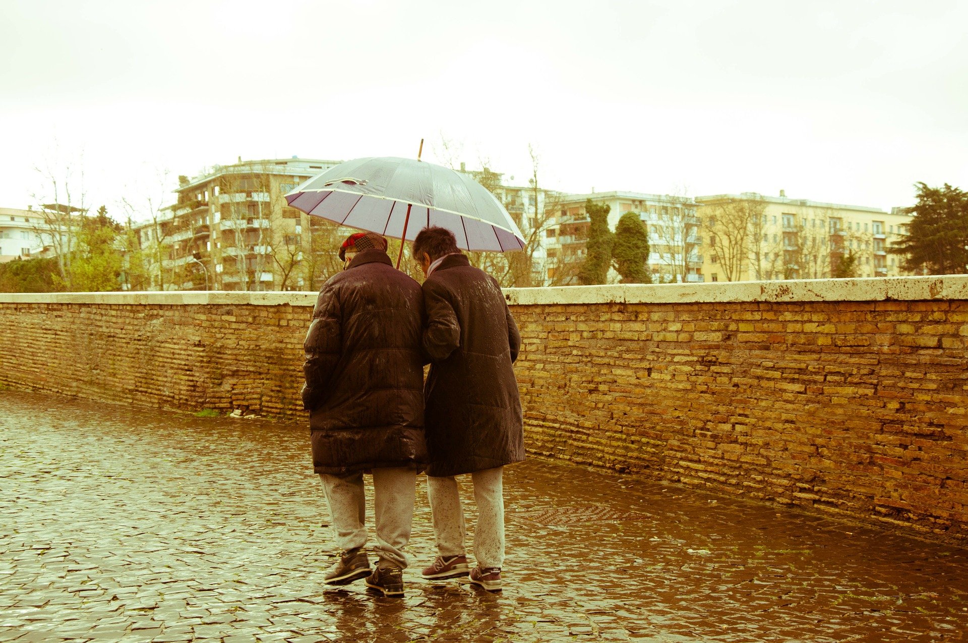 Casal de idosos caminhando em dia chuvoso, dividindo um guarda chuva