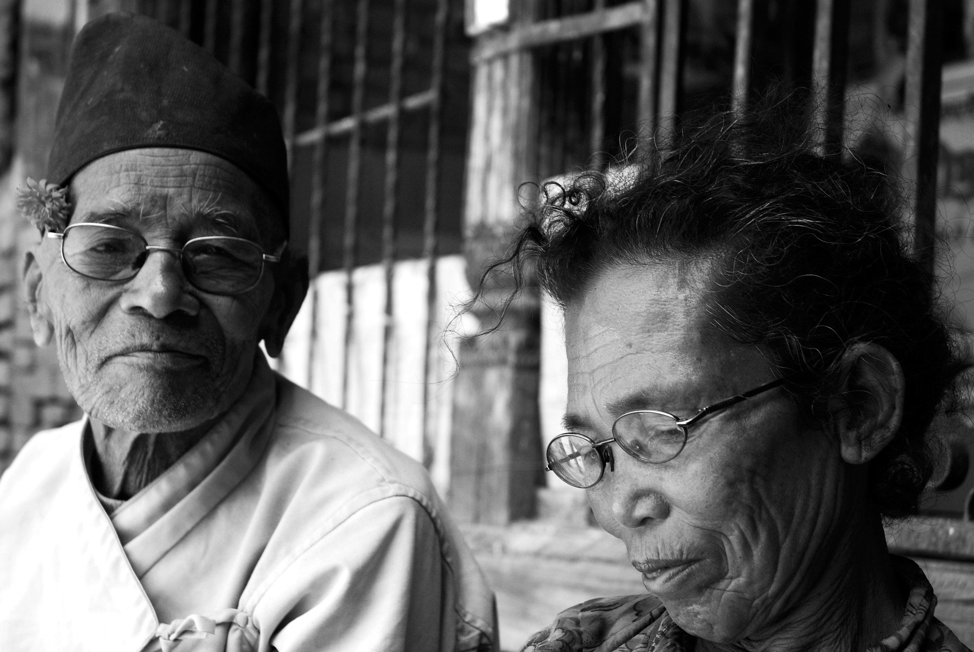 Foto presta e branca de casal de idosos orientais, ambos usam óculos e estão com uma expressão tentando segurar riso