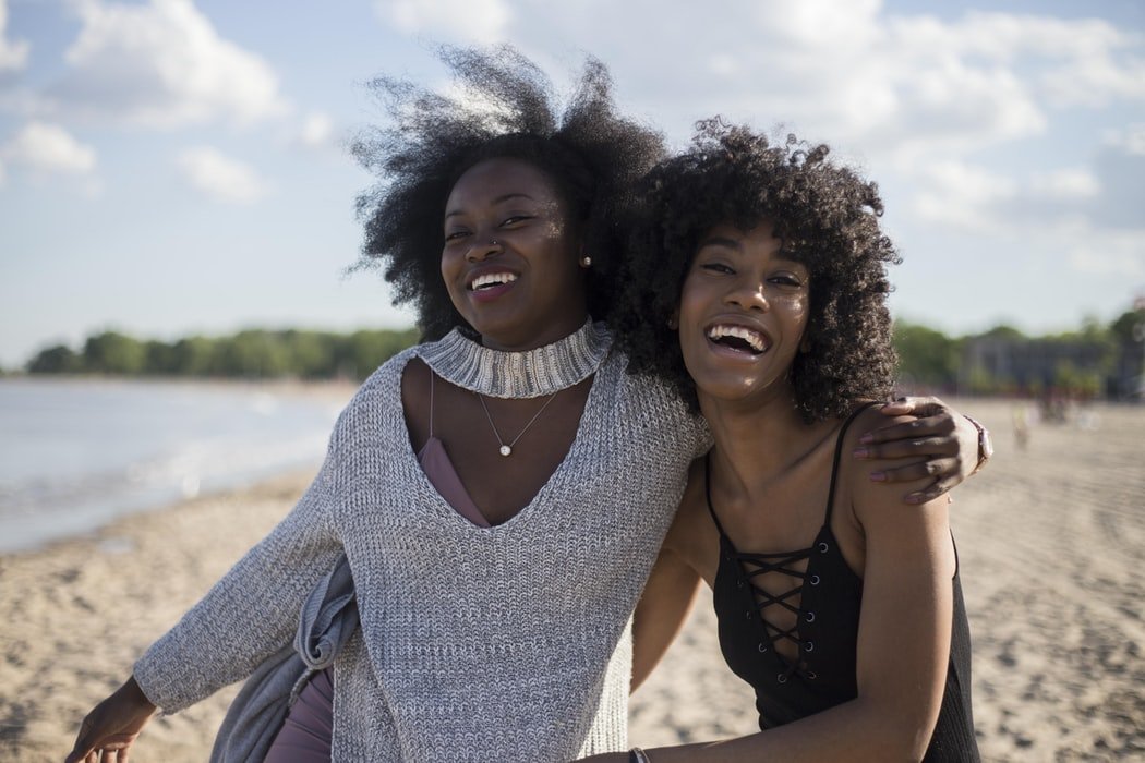 Duas mulheres negras, amigas se abraçando e sorrindo em praia