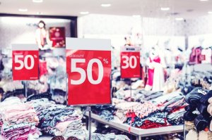 Pexels/Artem Beliaikin Mesas com pilhas de roupa dentro de loja, e em cada uma, uma placa anunciando um desconto como 50% e 30%.