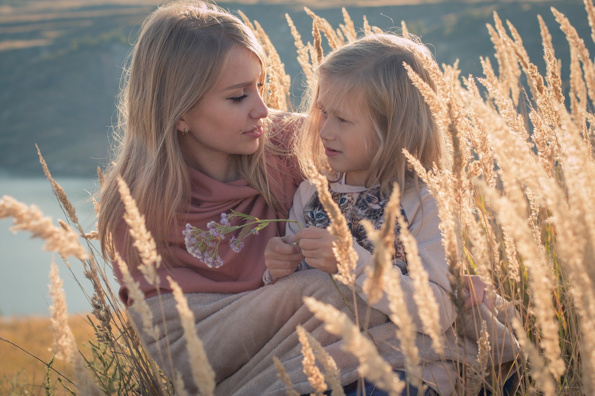 Mãe e filha enroladas em coberta, sentadas em campo de trigo