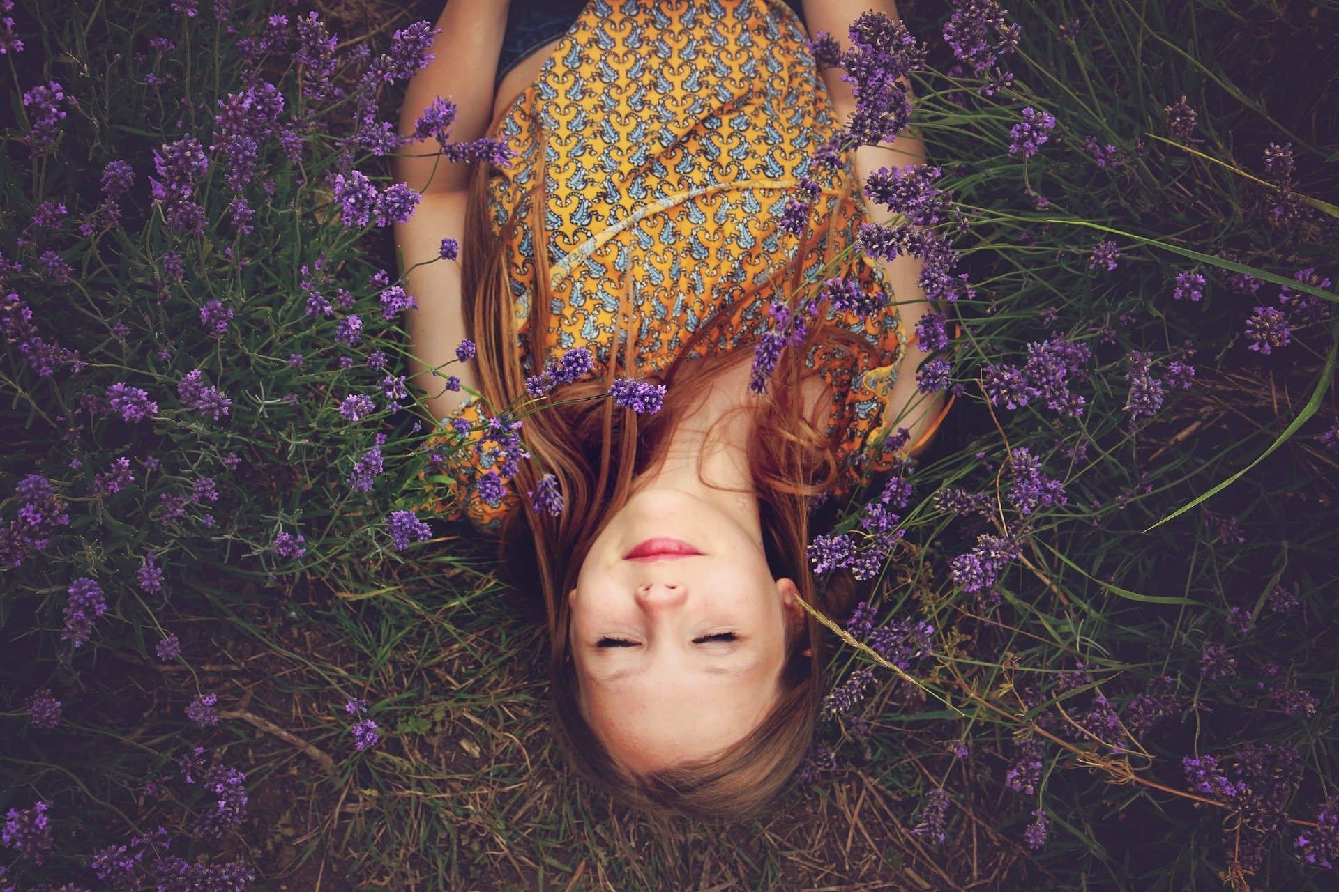 Menina deitada em campo, cercada de flores, de olhos fechados e sorriso no rosto