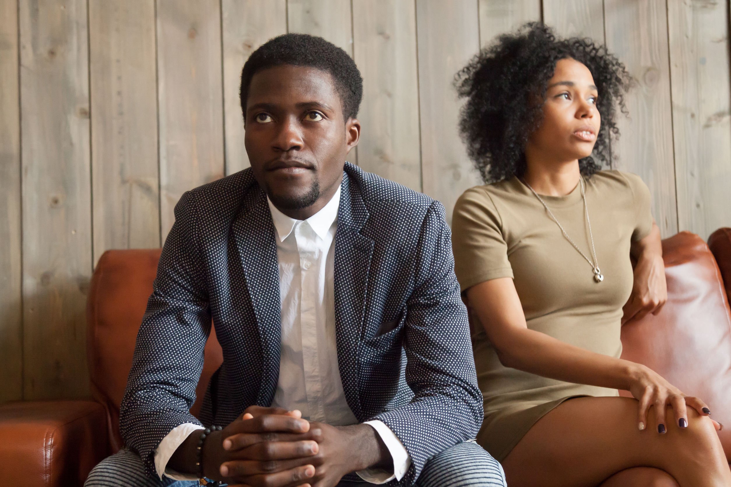 Homem e mulher, ambos negros, sentados lado a lado em sofá, porém olhando para lados opostos