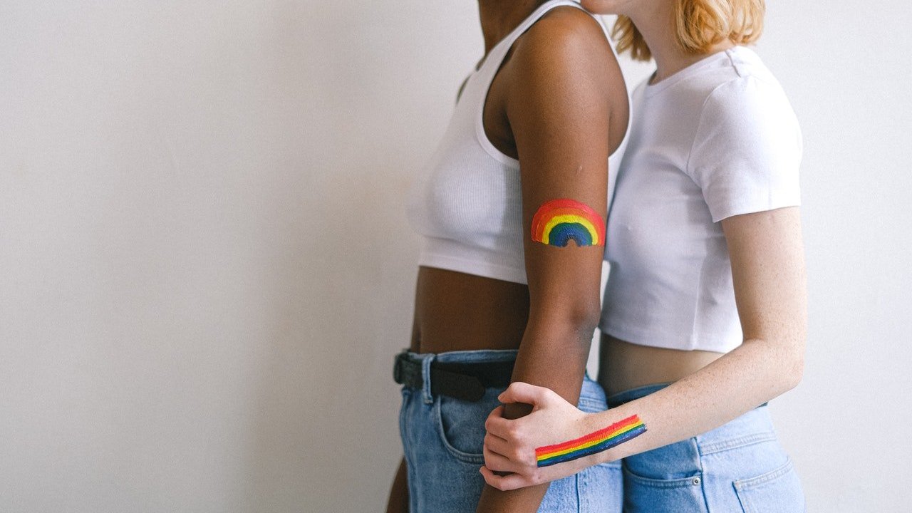 Duas mulheres abraçadas. Elas têm, pintados nos braços, símbolos LGBTQI+.